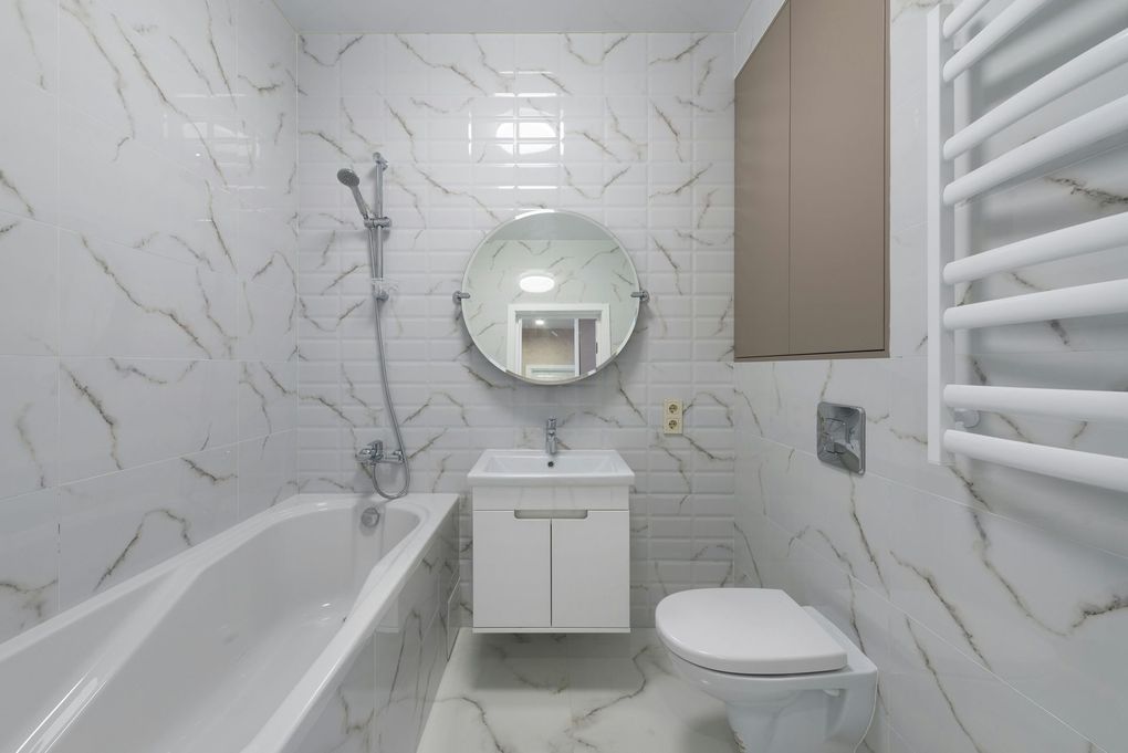 White bathroom with marble-look tiles, a bathtub, a sink, and a toilet. There's a round mirror and a towel warmer.