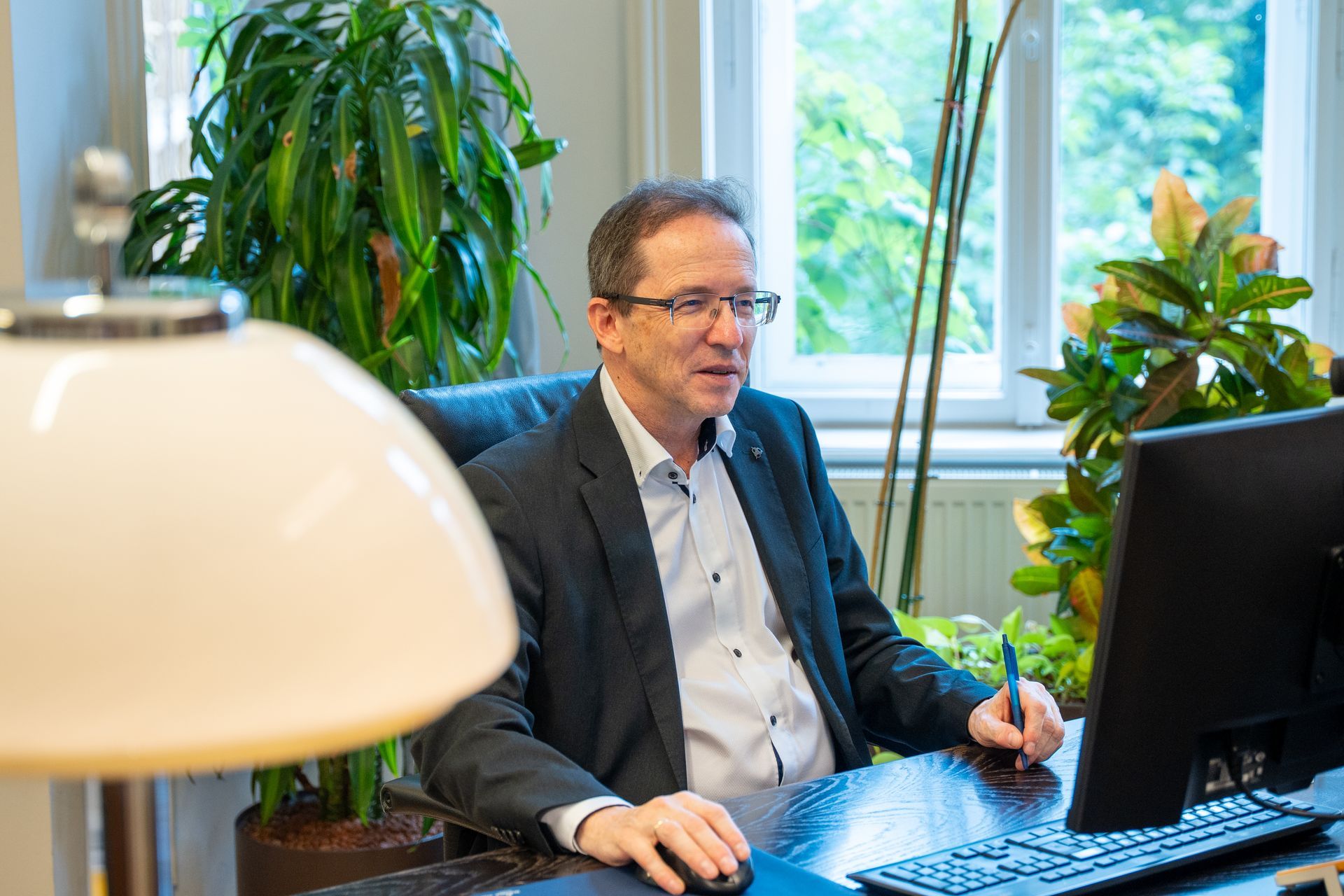 Ein Mann im Anzug sitzt an einem Schreibtisch mit einem Computer, umgeben von Pflanzen, und schaut auf den Bildschirm.