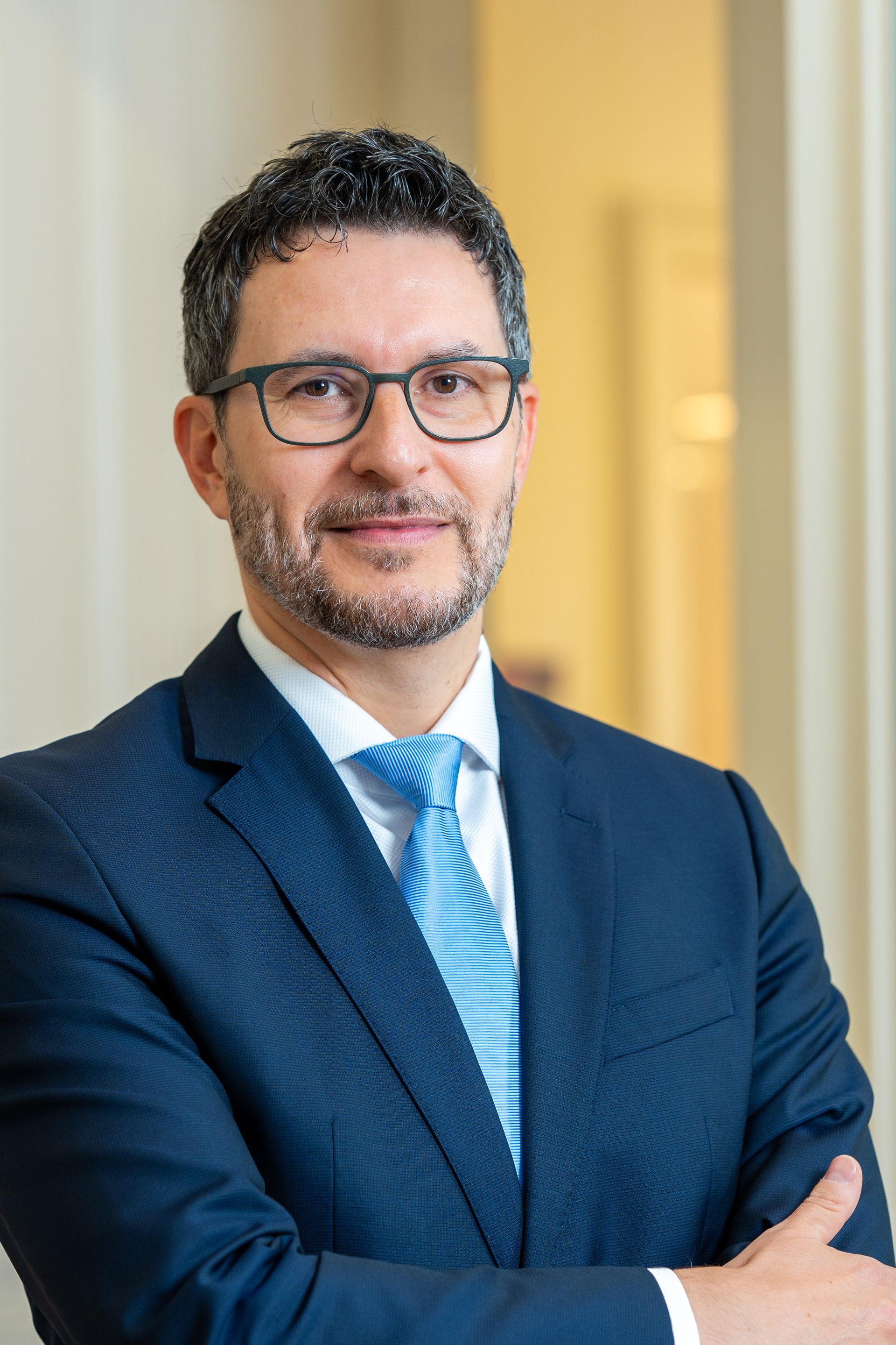 Mann mit Brille und blauem Anzug, die Arme verschränkt, lächelt in einem Büro.