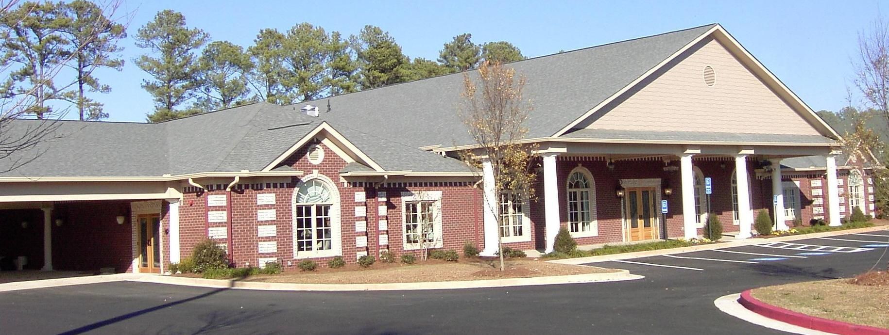 Single-story brick building with white columns and a covered entrance, set beside a parking lot and trees.