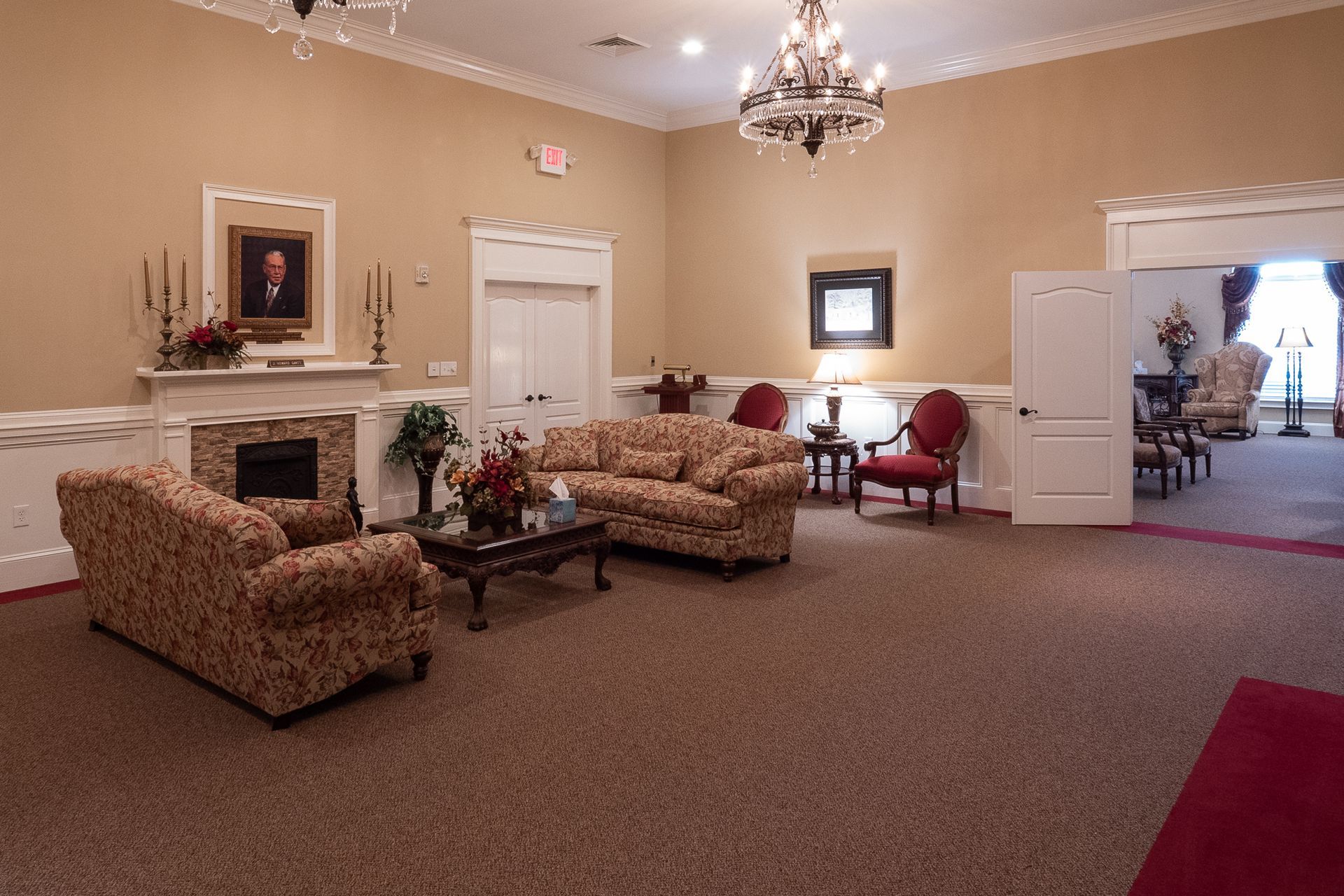 Elegant lounge with patterned sofas, chandelier, fireplace, and an open doorway into another room.
