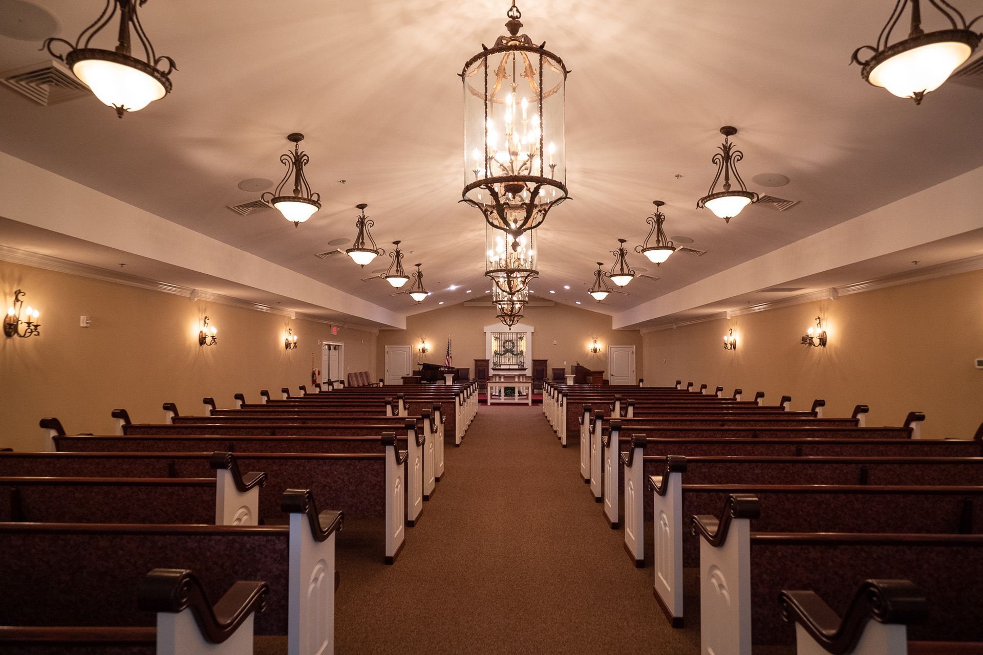 Symmetrical church interior with wooden pews, central aisle, and hanging chandeliers under warm lighting