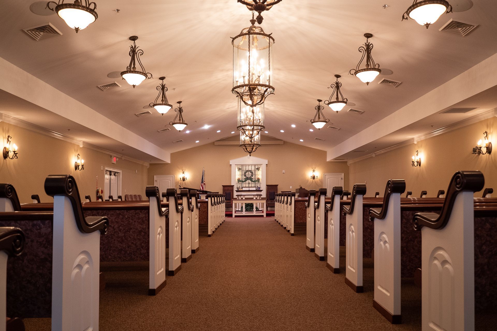 Interior of a quiet chapel with rows of pews, chandeliers, and a central aisle leading to an altar