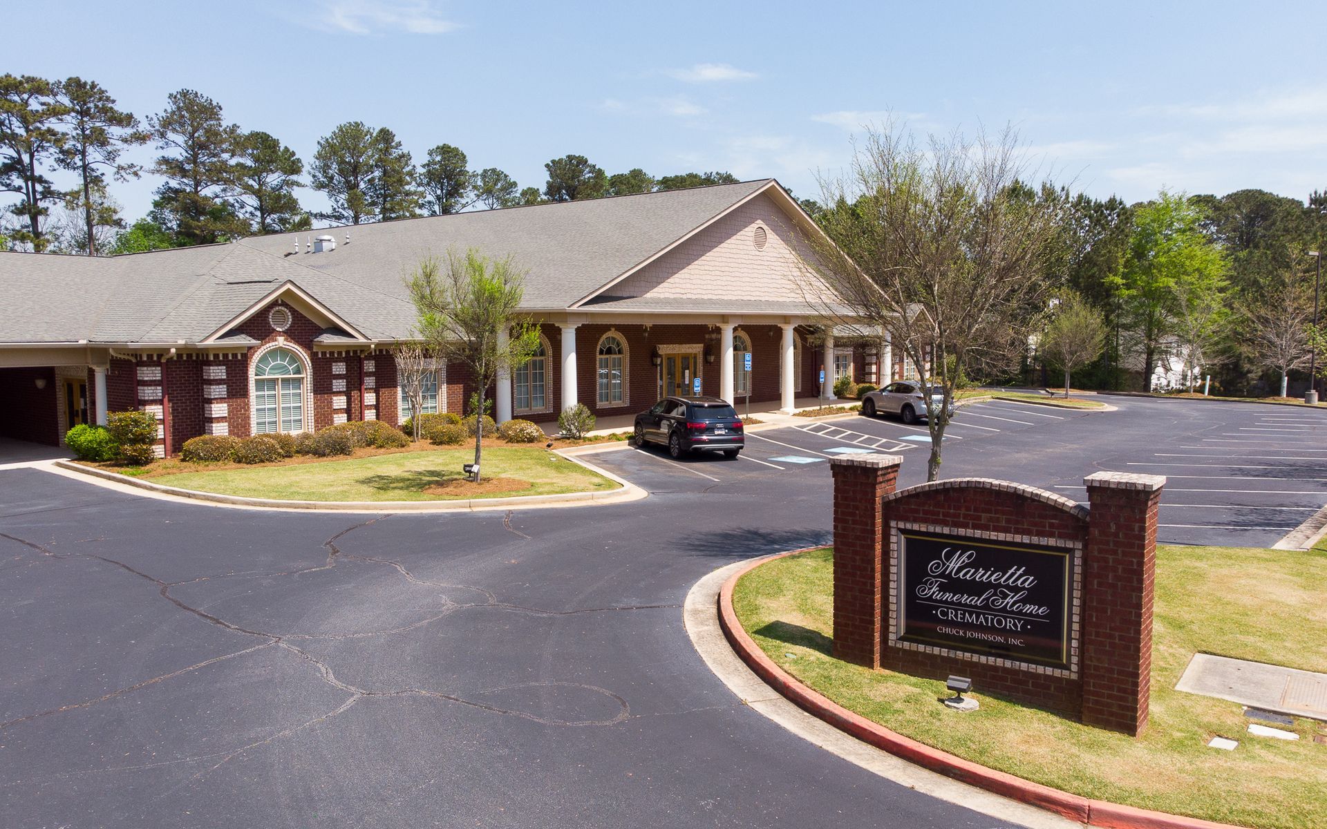 Brick building with a covered entrance and parking lot, viewed from a driveway with a sign in front.