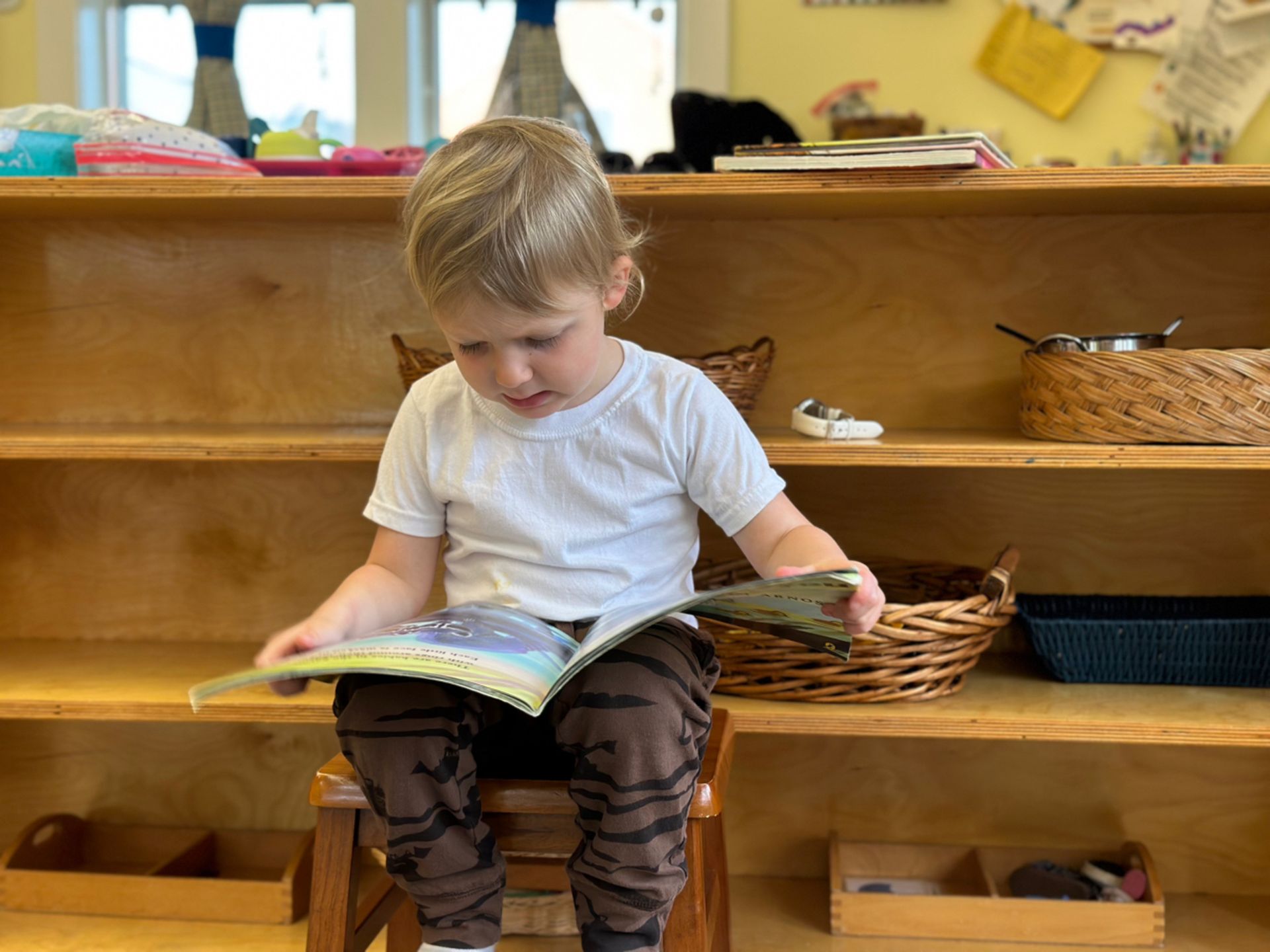 A Montessori young boy sits on a stool reading a book