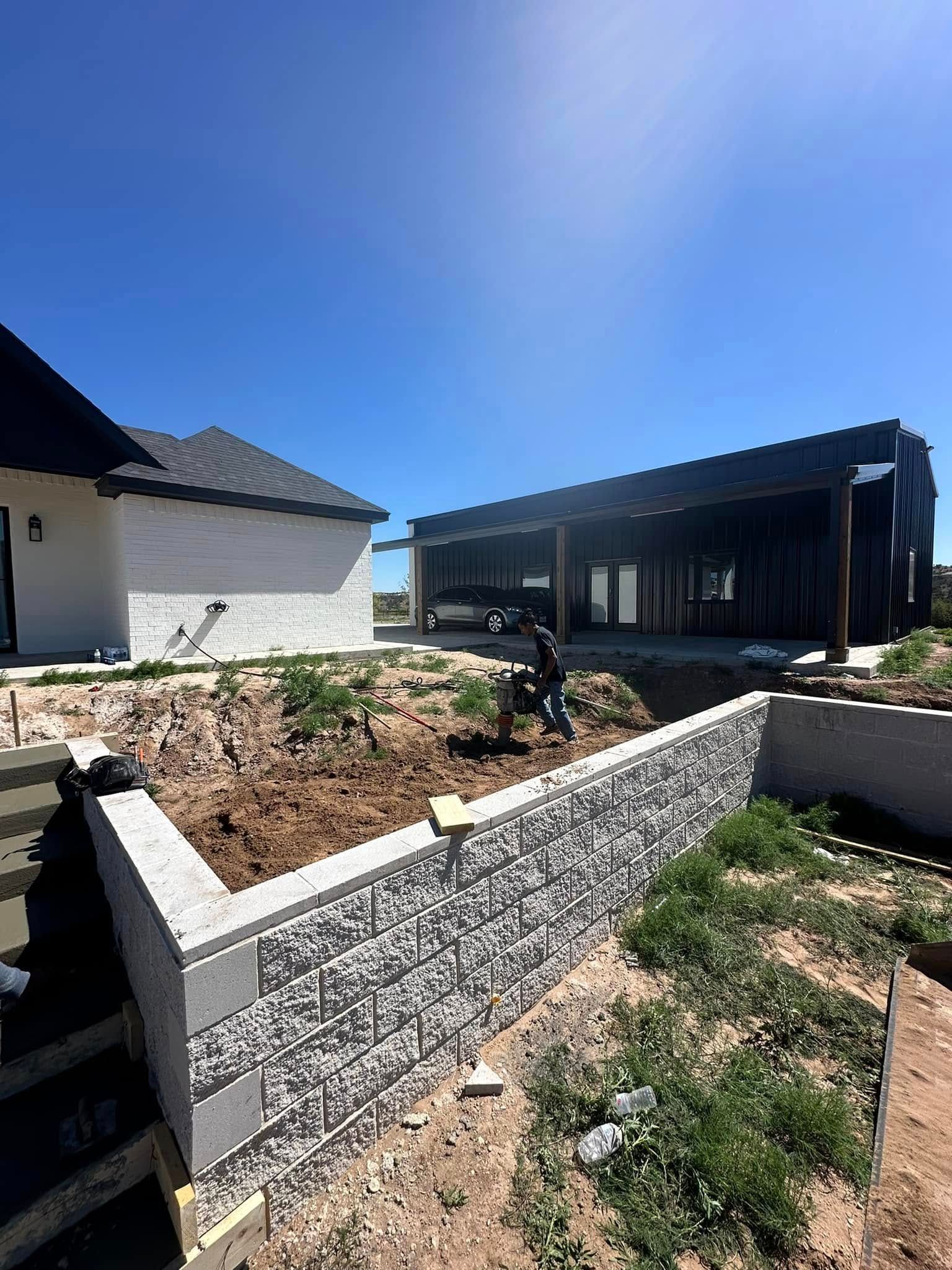 New home construction beside a white house, with a concrete block retaining wall under a clear blue sky