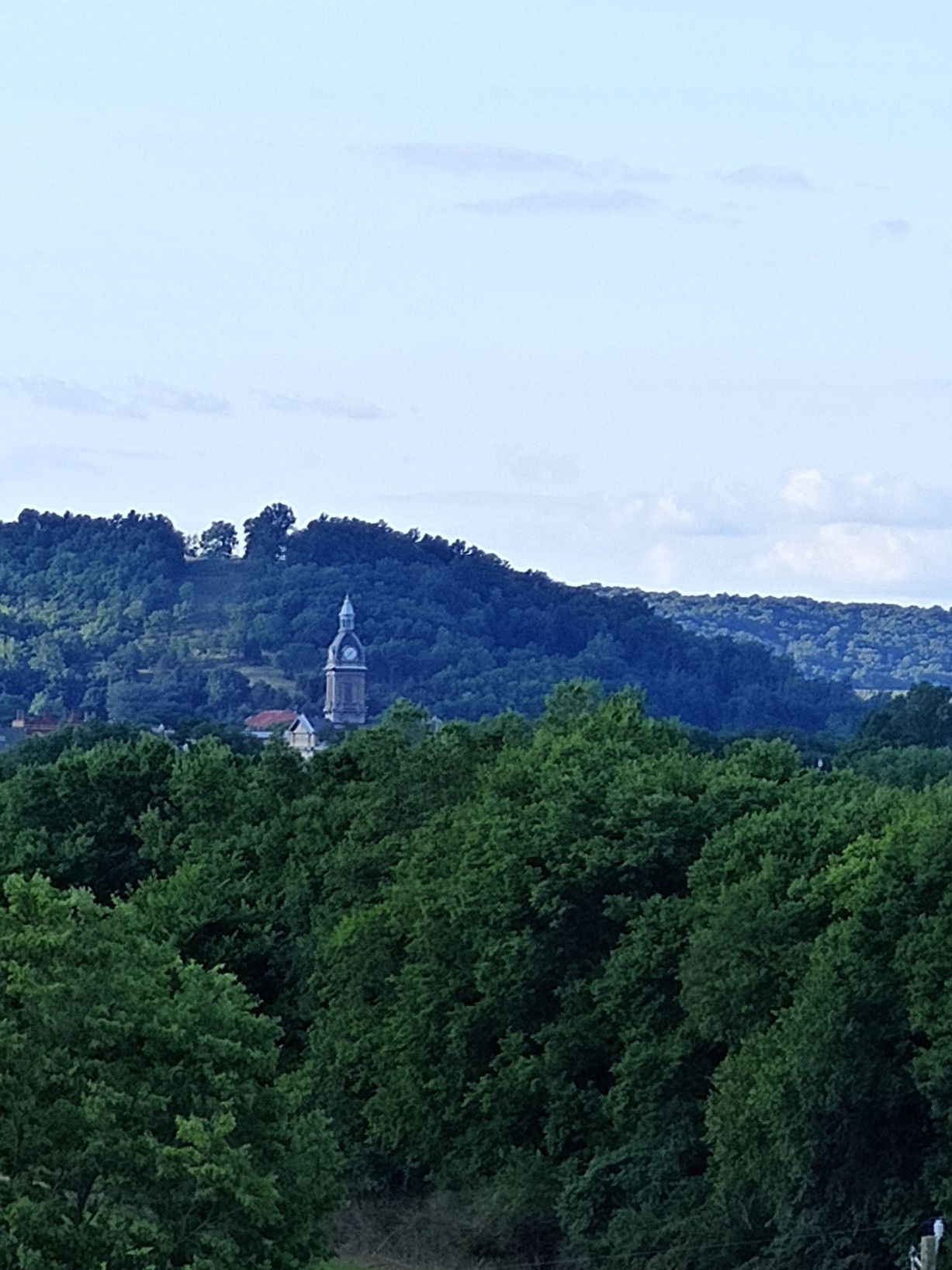 View of a clock tower atop a forested hill, green trees in the foreground, under a cloudy sky.