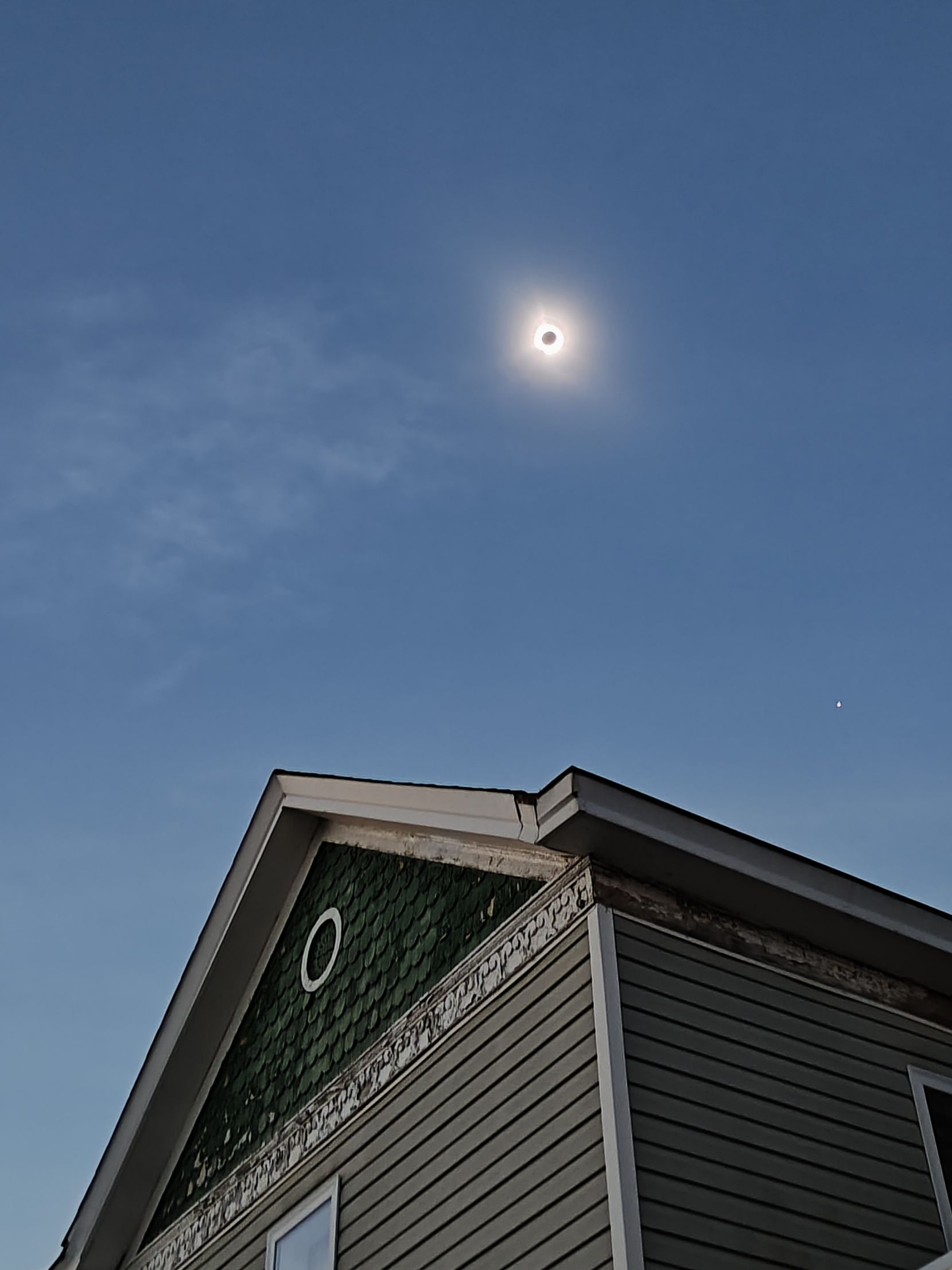 Solar eclipse in a blue sky above a house with green roof shingles and siding.