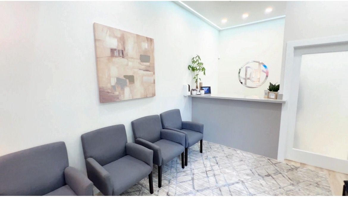 Reception area with chairs, desk, plants, and artwork. Neutral colors, natural light, and patterned carpet.