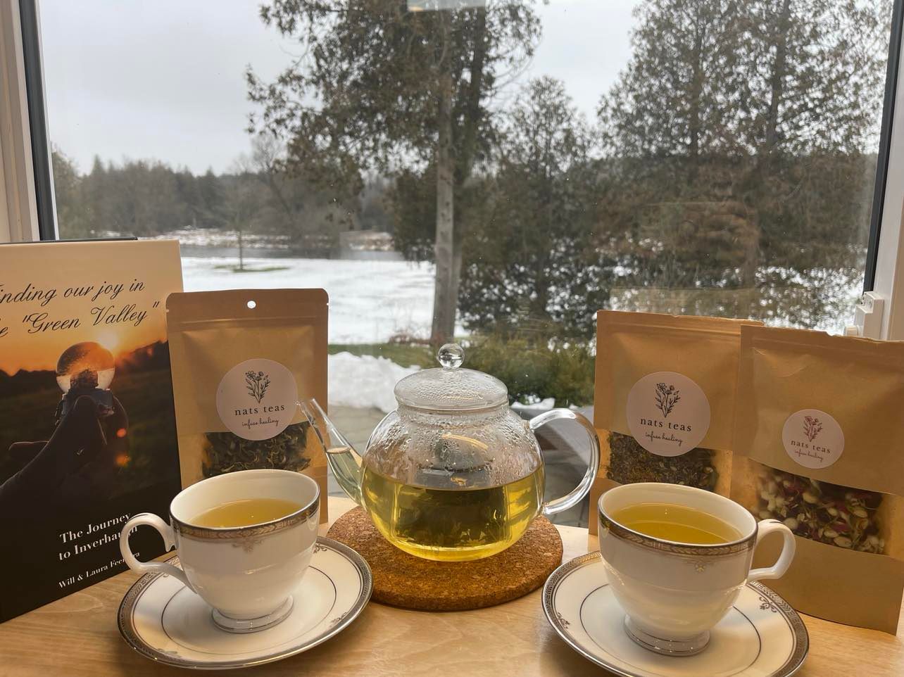 Tea set with a view: cups, teapot, and tea bags in front of a snowy landscape seen through a window.
