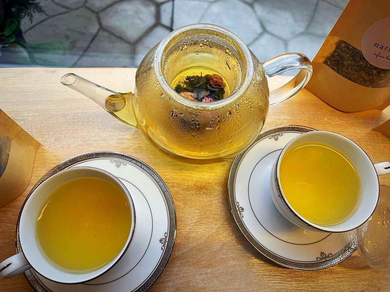 A glass teapot and two teacups filled with golden tea sit on a wooden surface.