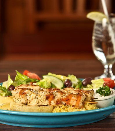 Grilled chicken skewer on a plate with salad, olives, and dipping sauce; glass of water.