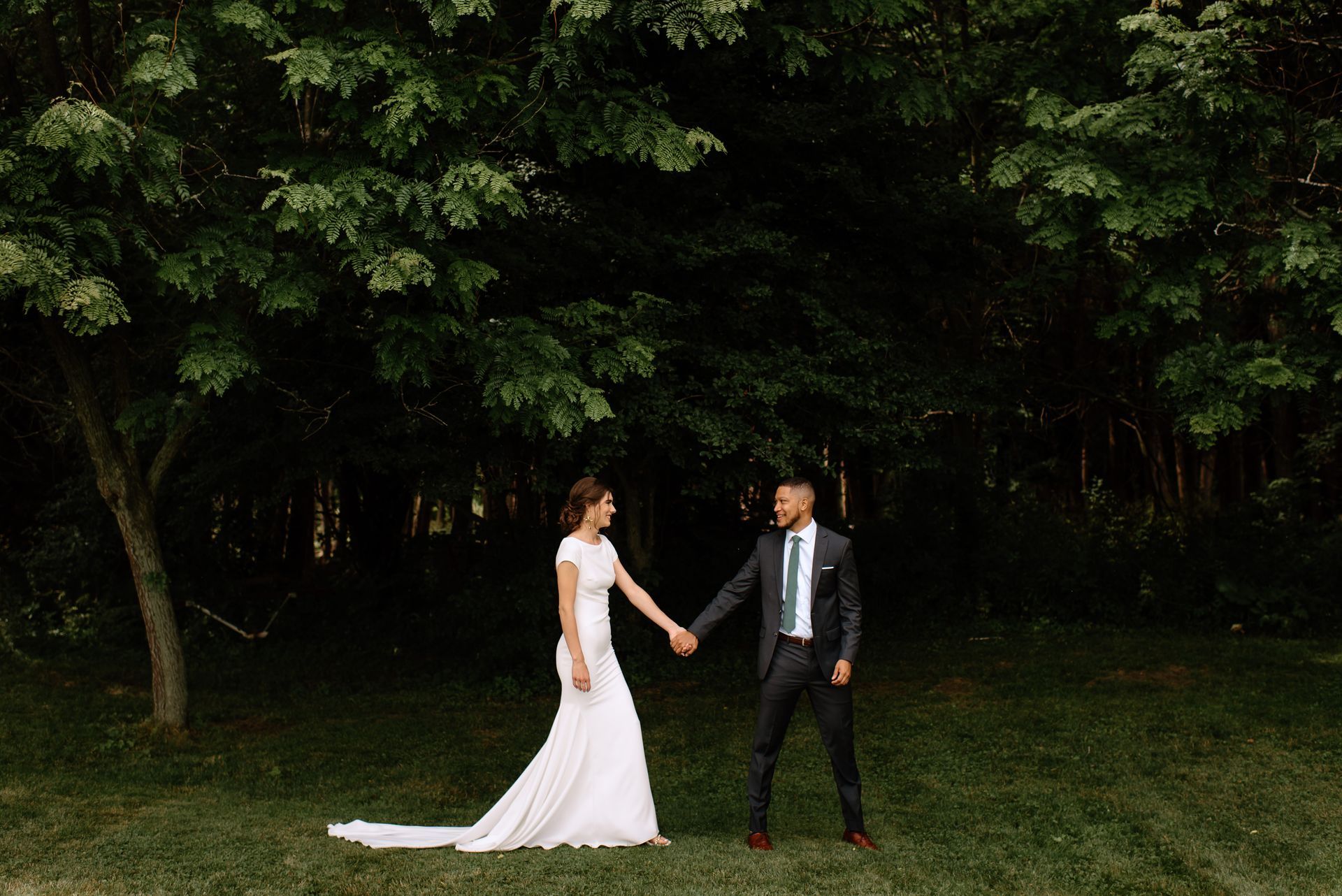 Bride and groom holding hands, standing in a forest. The bride wears a white gown, the groom wears a dark suit.