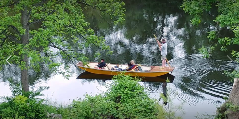 People on a wooden boat in a river, surrounded by lush green trees. One person is standing with arms raised.