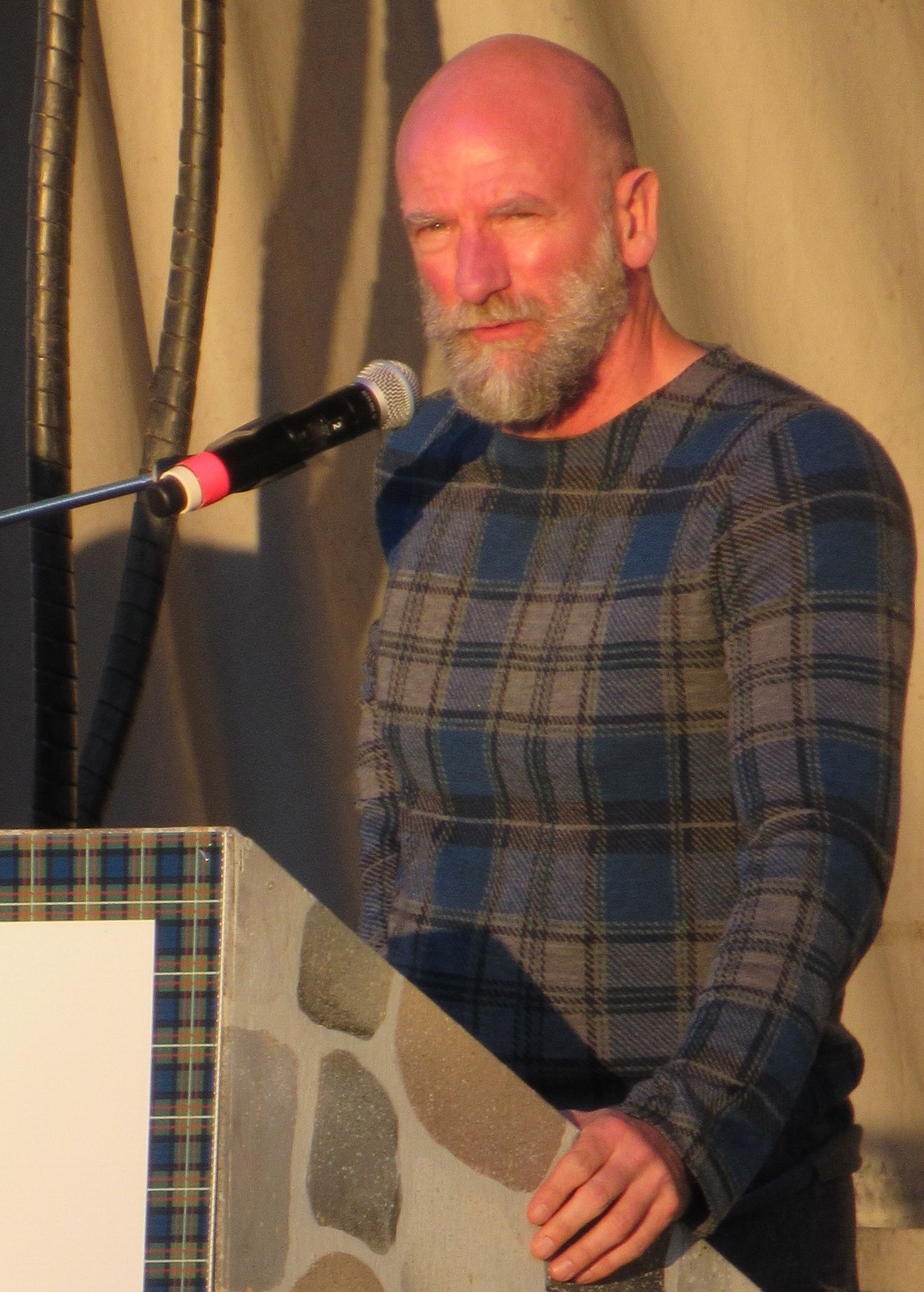Bald man with beard speaking at a podium; wearing a blue plaid sweater, microphone.