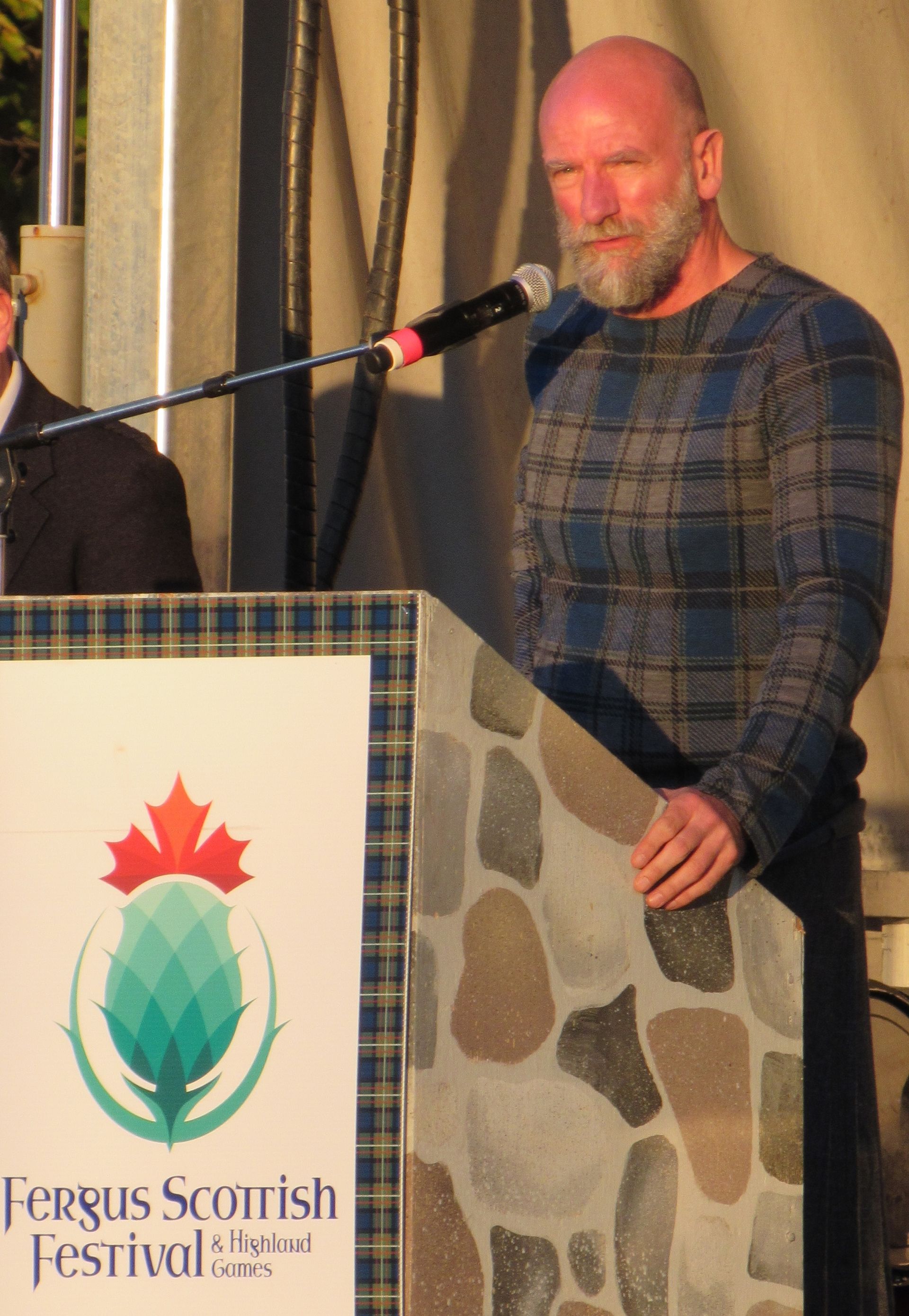 Man in plaid shirt speaks at a podium with Fergus Scottish Festival logo.