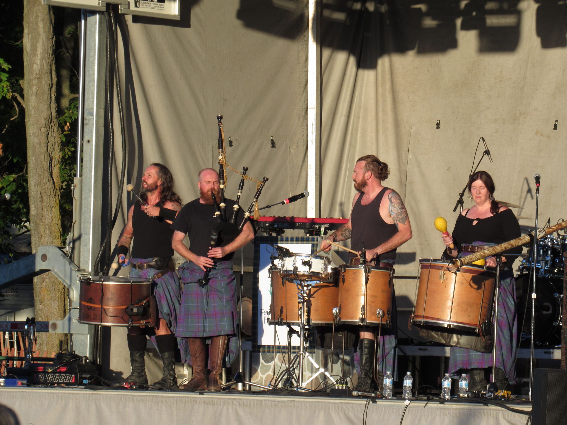 Scottish band on stage: bagpipes, drums, kilts. Musicians perform with focused expressions, in natural light.