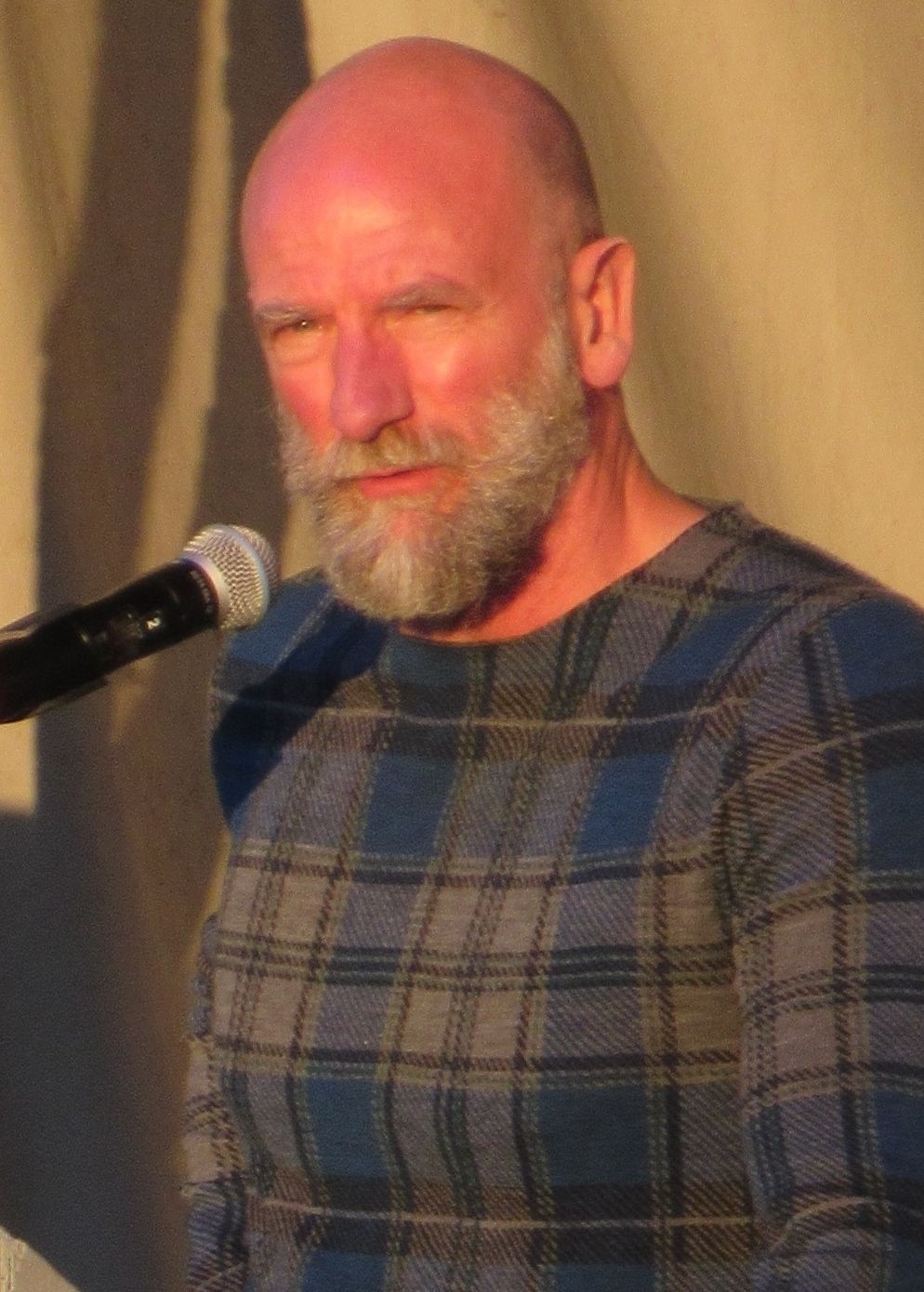 Bald man with gray beard, in blue plaid sweater, speaking into a microphone.