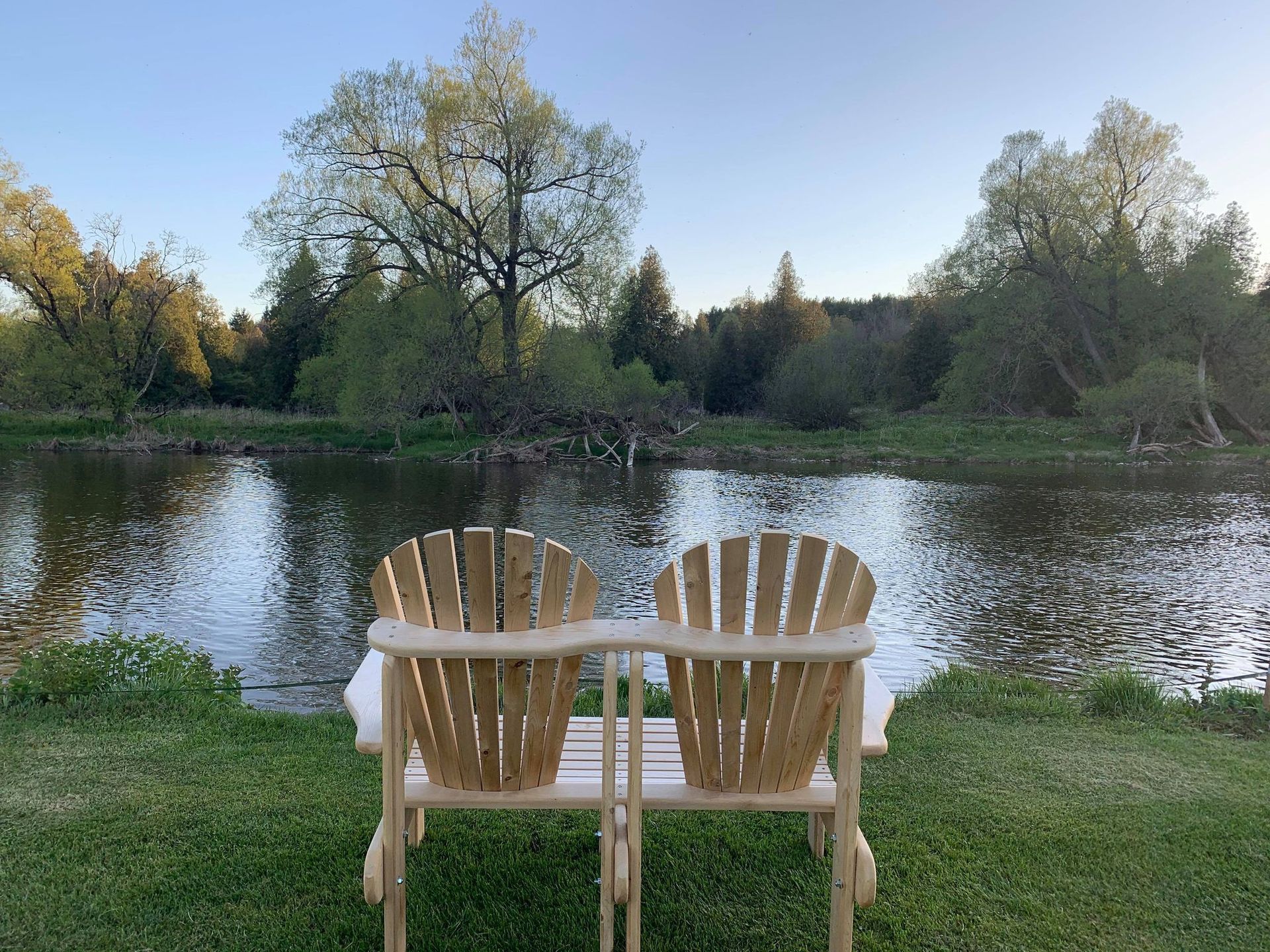 Two wooden chairs sit on a grassy bank, facing a calm river reflecting trees, in a serene outdoor setting.