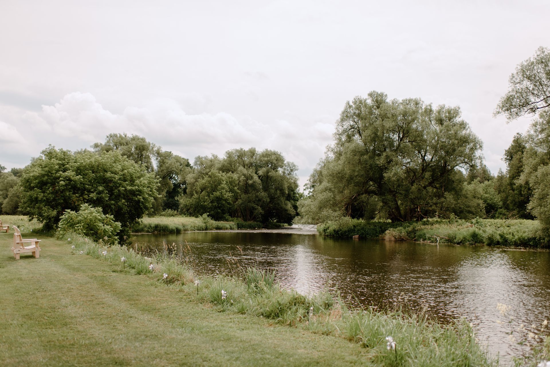 A calm river flows past a grassy bank, with trees lining the water under a cloudy sky.