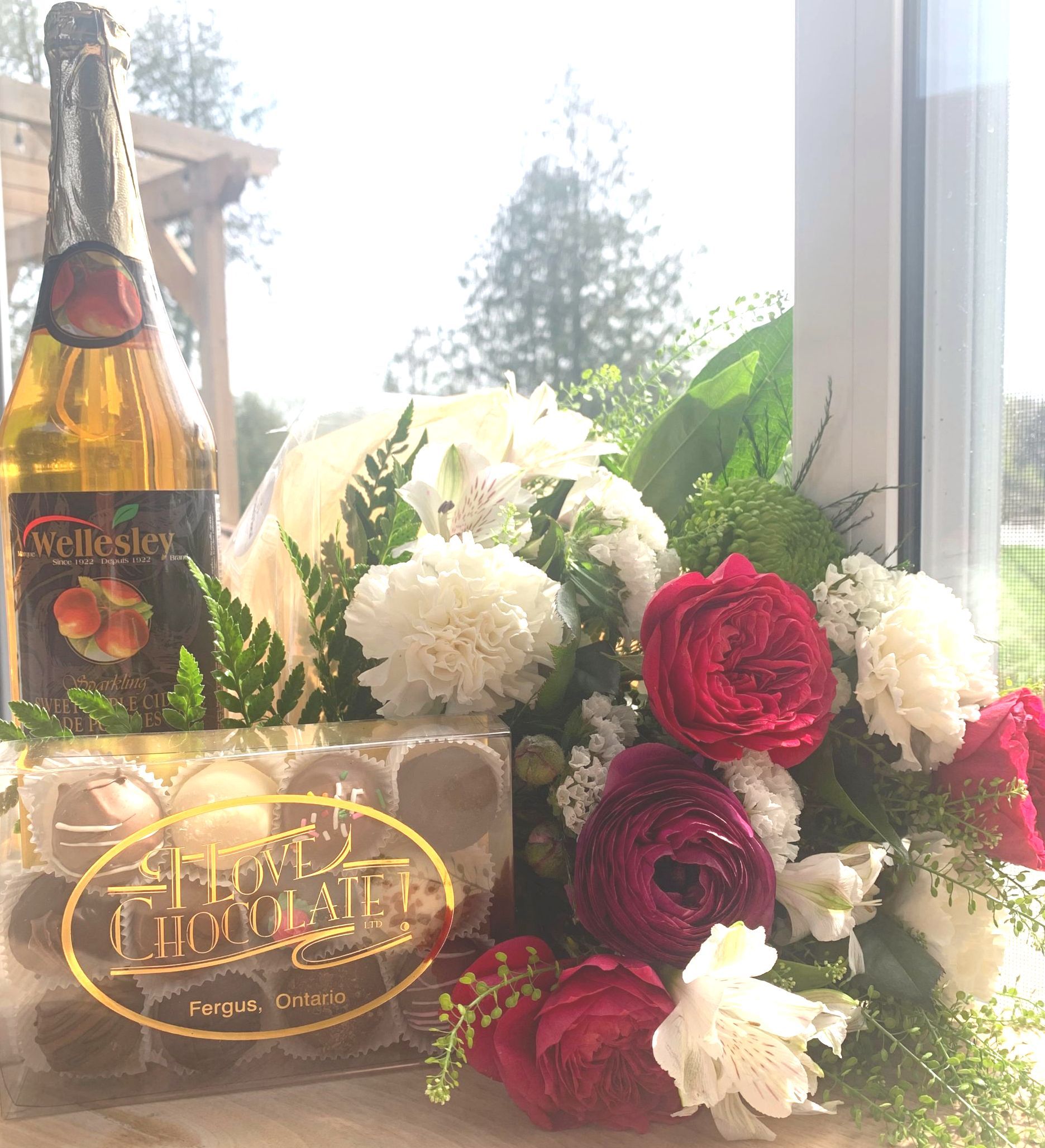 Champagne bottle, chocolates, and flowers in front of a window. Red and white floral arrangement.
