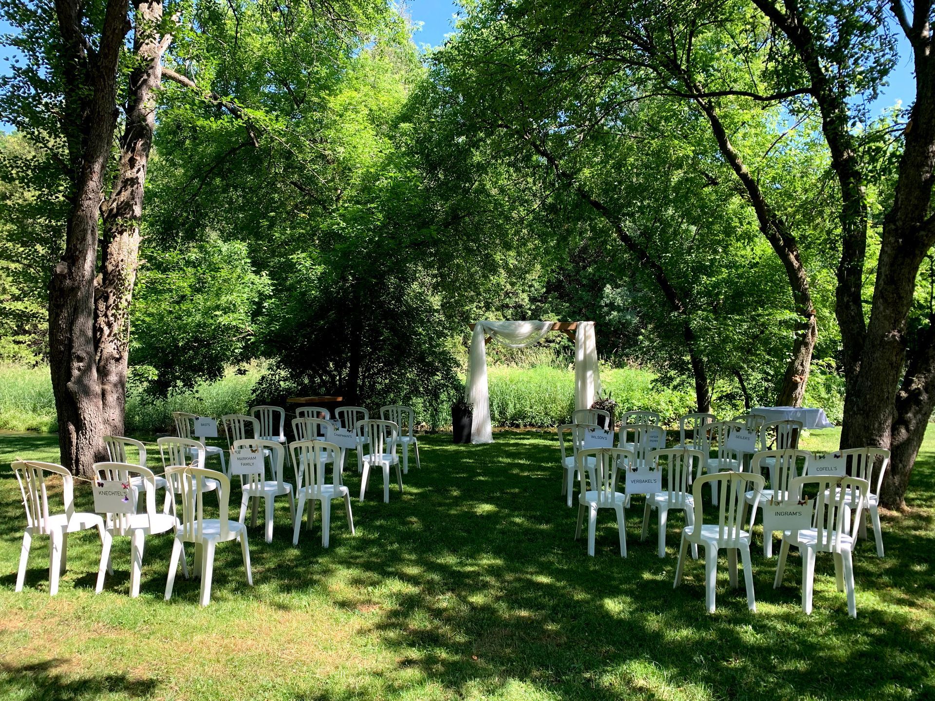 Outdoor wedding ceremony with white chairs arranged on grass under trees.