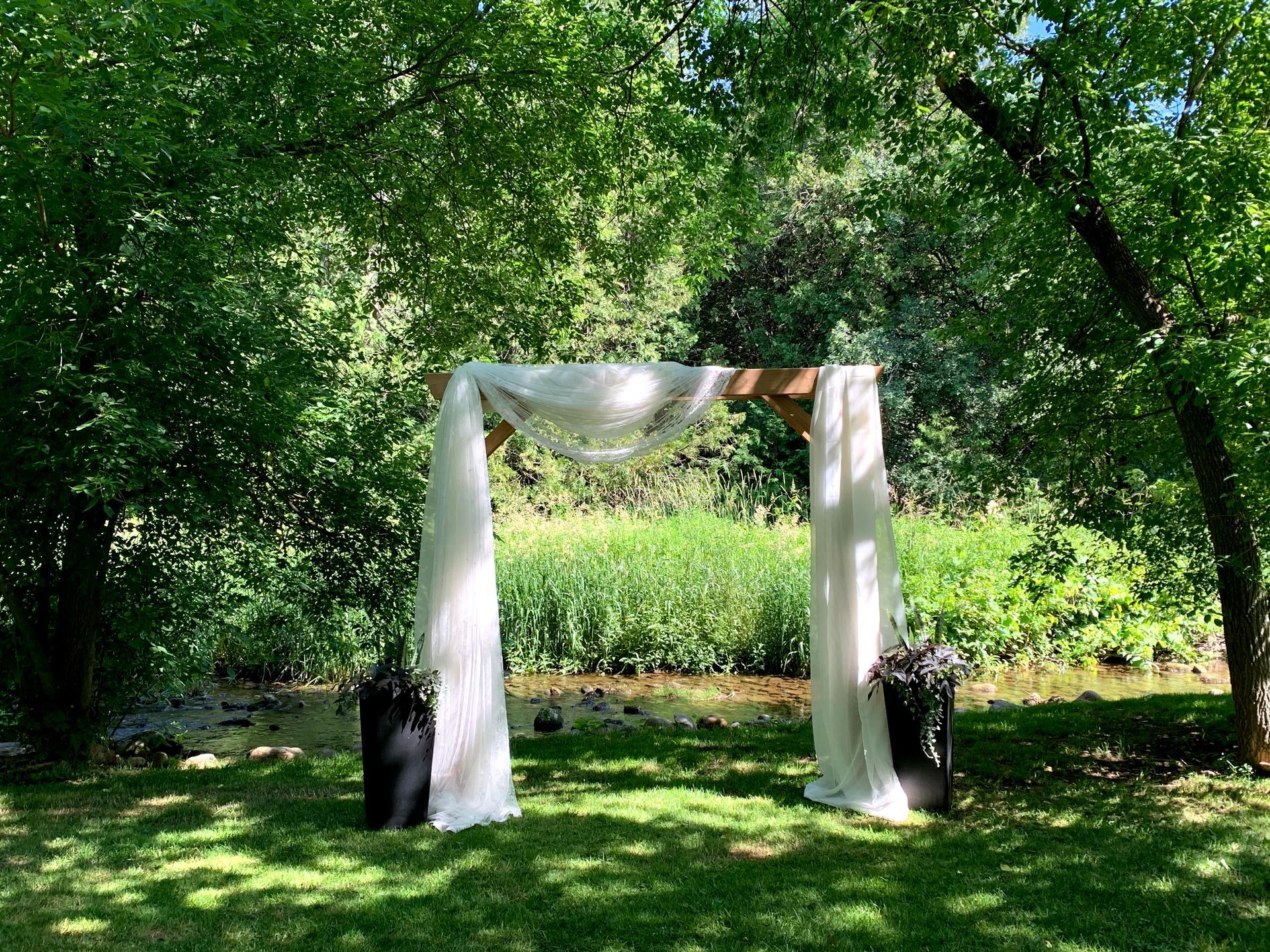Wedding arch with draped fabric and flowers in a wooded outdoor setting.