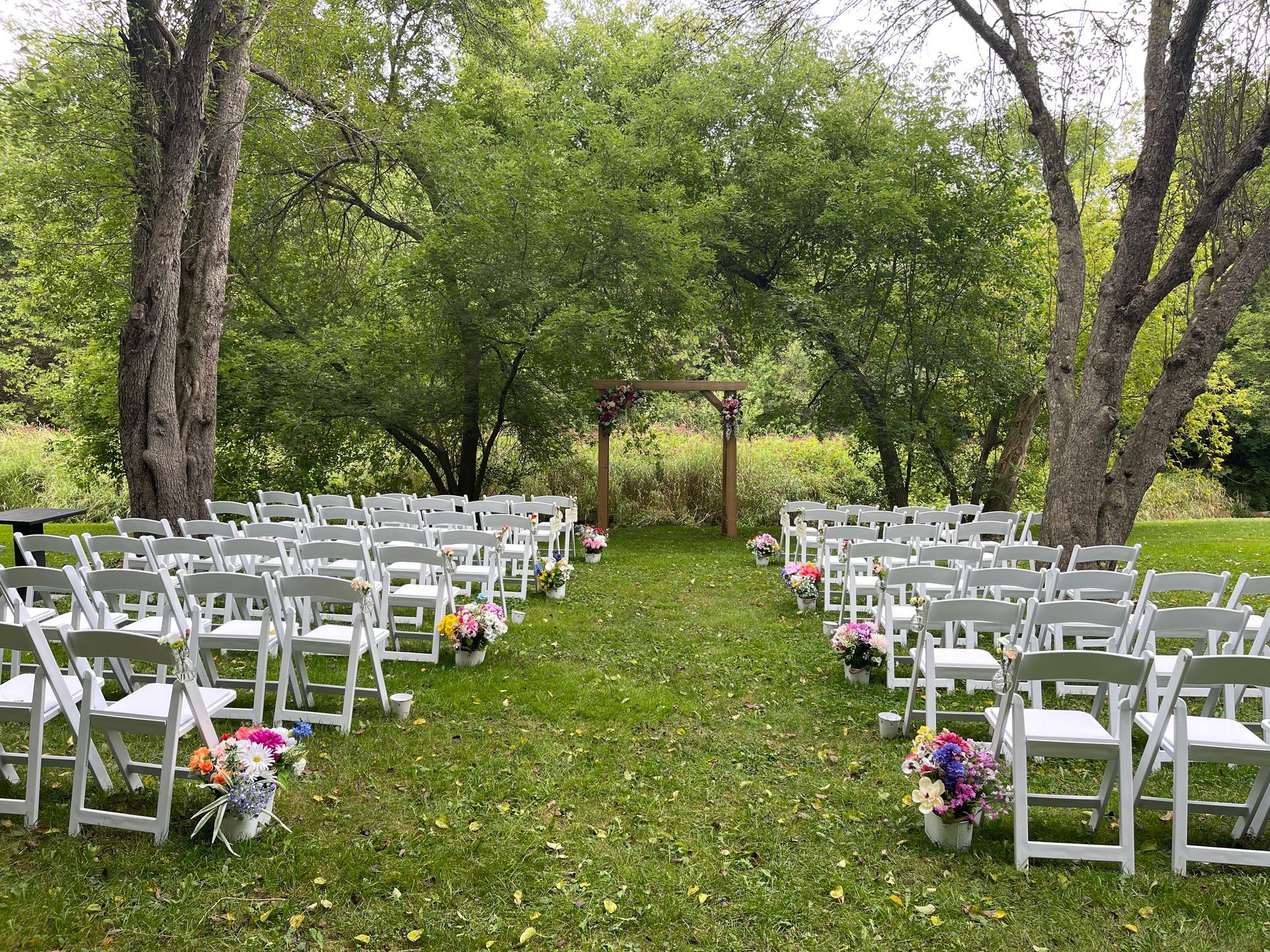 Outdoor wedding aisle with white chairs, flower arrangements, and a wooden arch in a green garden