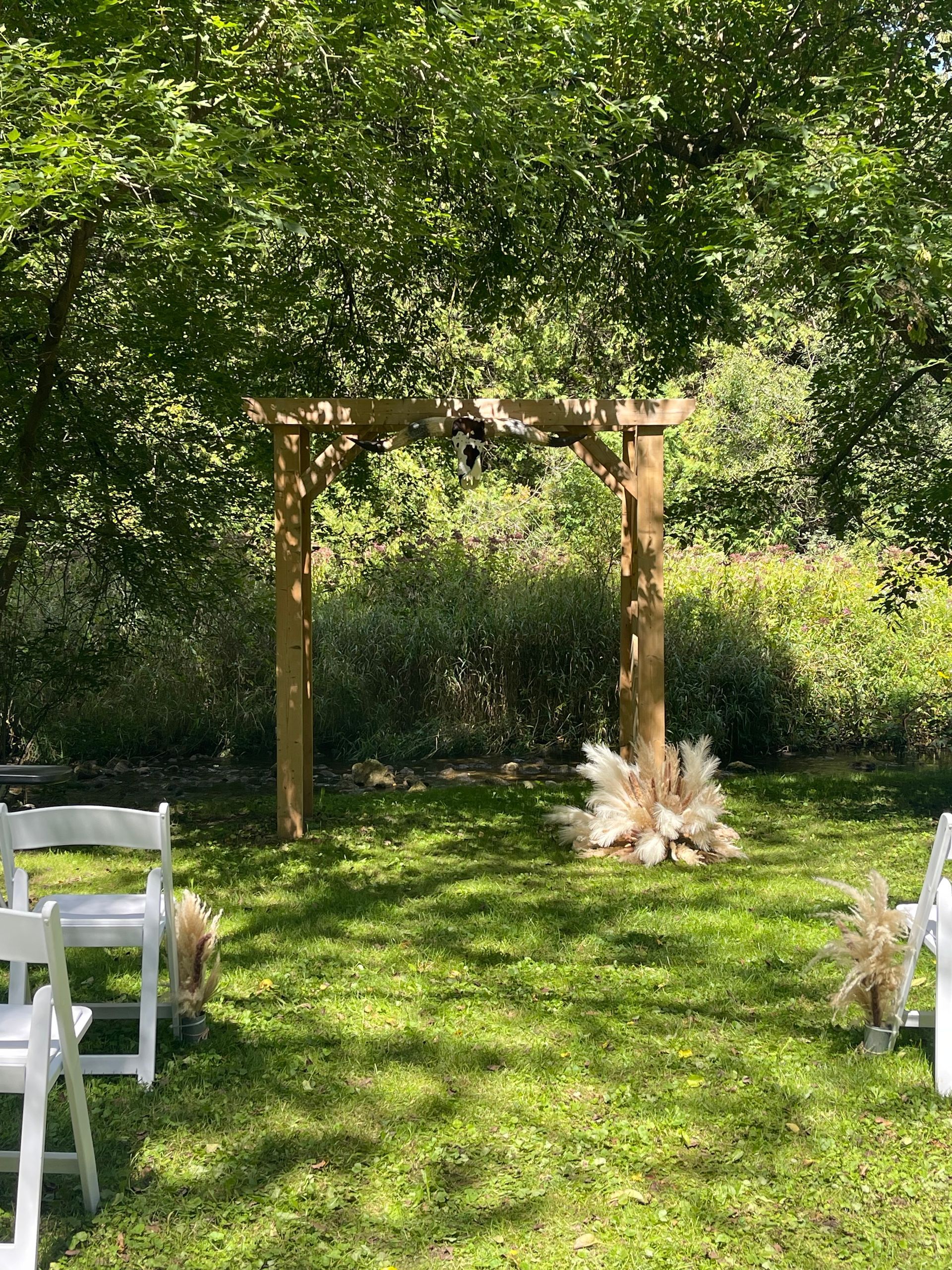 Wooden archway in a sunny garden with white chairs and floral decorations on the grass.