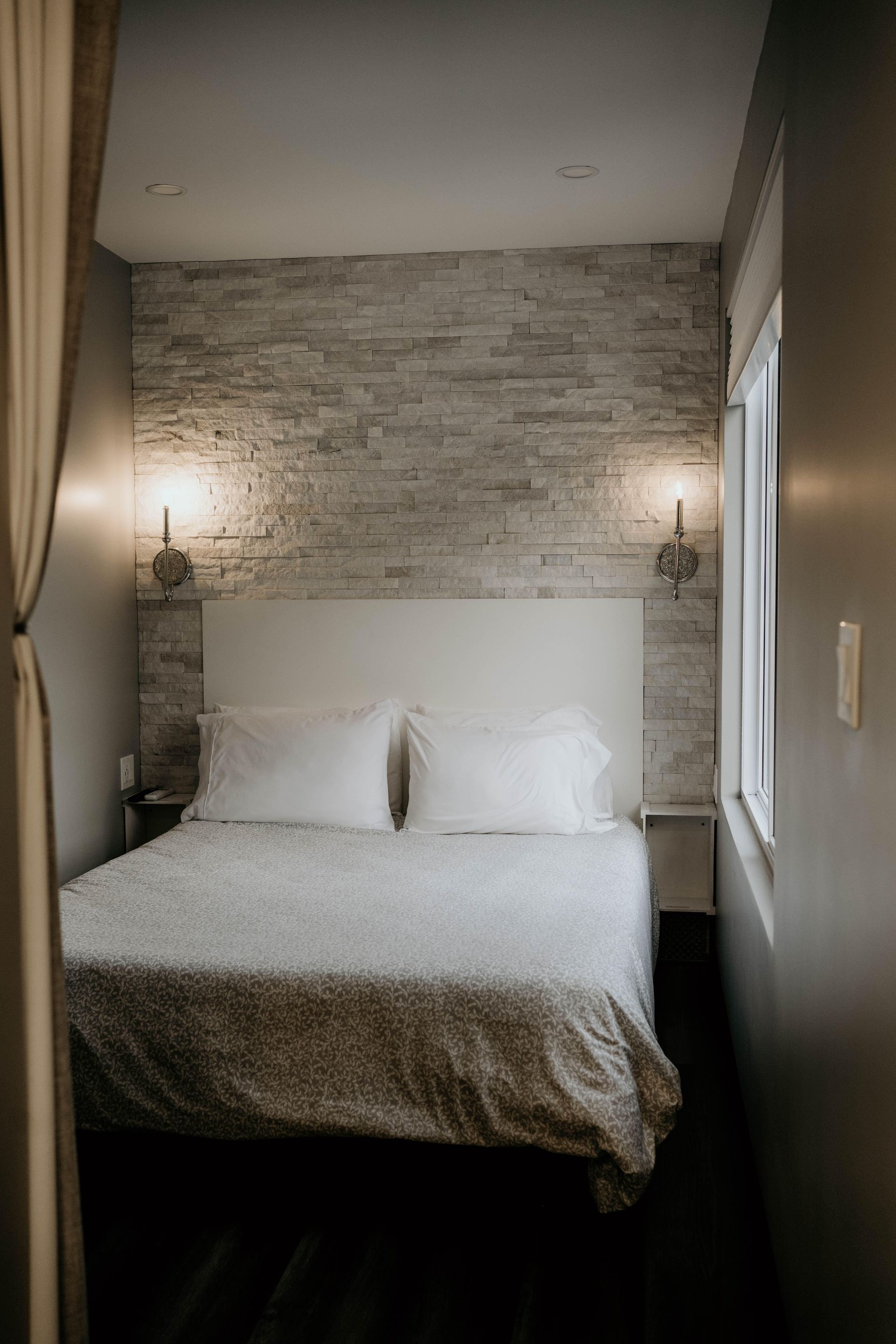 Cozy bedroom with a white bed, stone accent wall, sconce lighting, and a window.