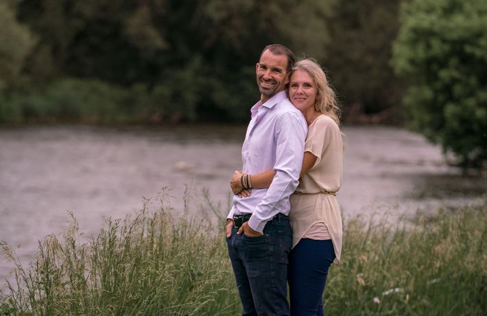 Couple embracing by a river; man smiling, woman resting head on his shoulder, natural setting.