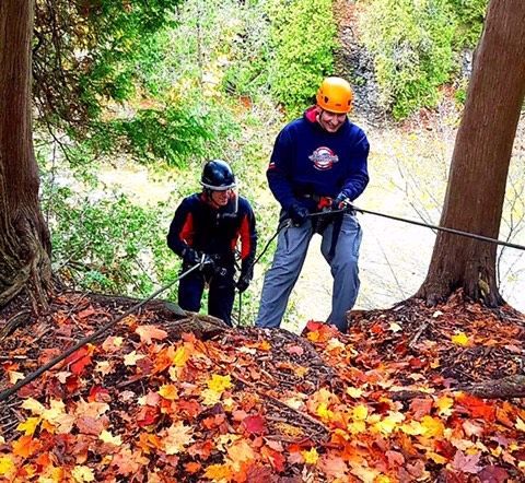 Two people rappelling down, surrounded by autumn leaves. One wears orange helmet, the other a black one.
