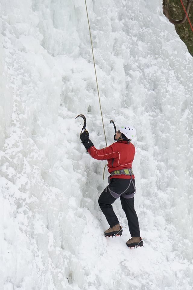 Ice climber in red jacket and helmet scaling a frozen waterfall, using ice axes and rope.
