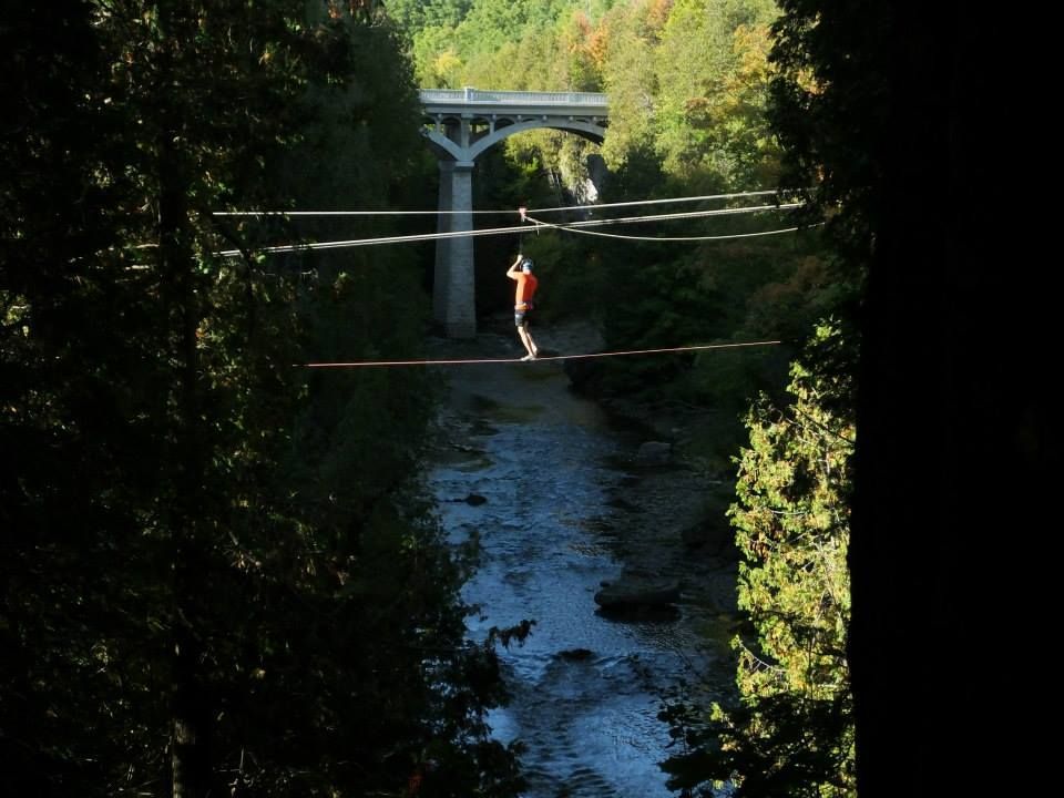 Person walking a slackline over a river with bridge in the background, framed by trees.