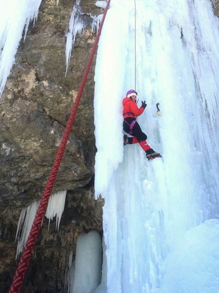Ice climber in red jacket ascends a frozen waterfall, with red rope and ice tools visible.