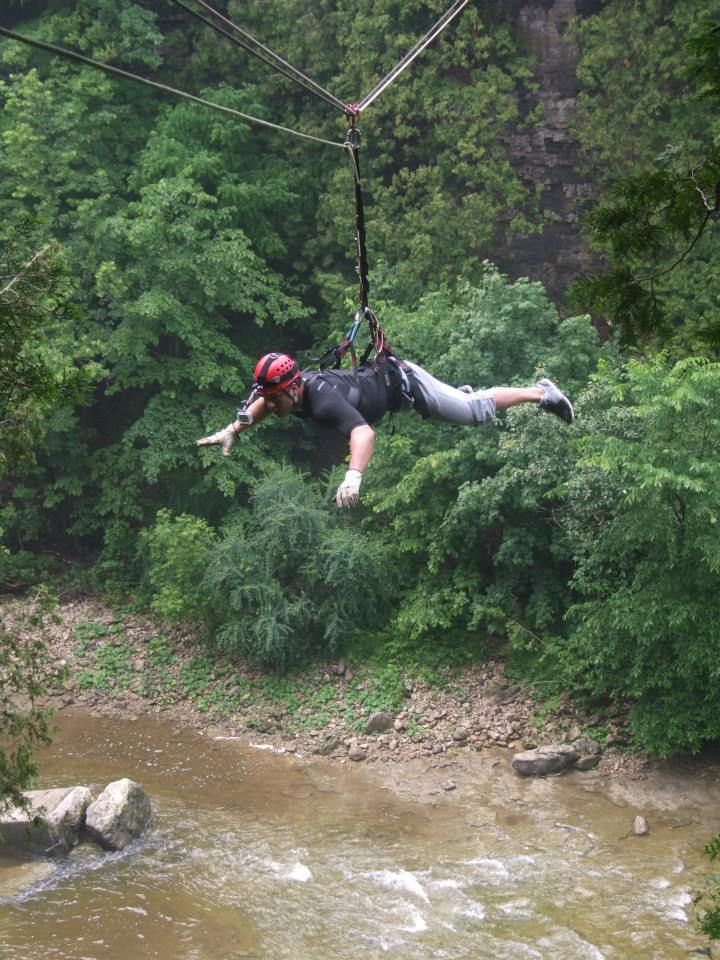Man ziplining over a river, arms outstretched, wearing helmet and harness, surrounded by green trees.