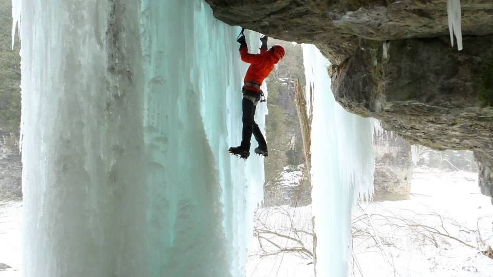 Ice climber in red jacket, hanging from a rock overhang with ice formations.