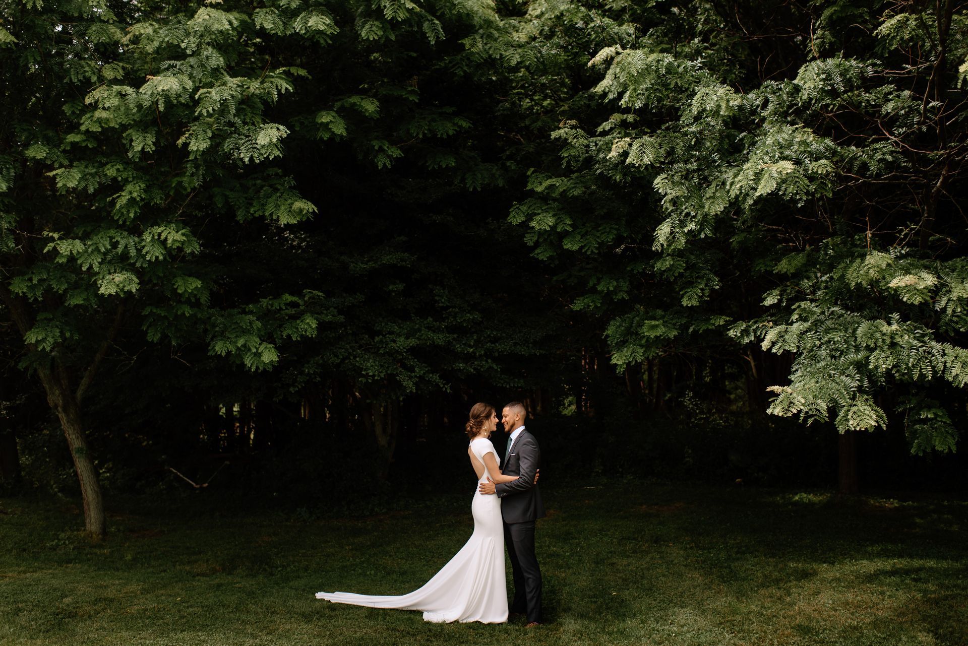 Bride and groom embrace under lush green trees. Woman in white dress, man in gray suit.