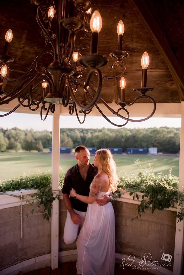 Couple embraces in a gazebo, lit by a chandelier, overlooking a field.