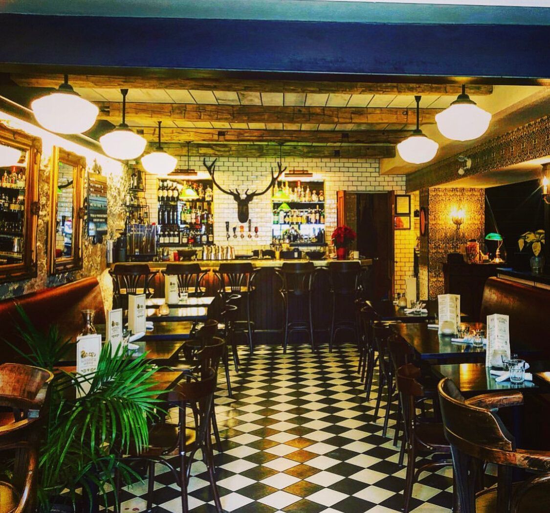 Interior of a restaurant, black and white checkered floor, dark wooden tables, bar, stag head on the wall, overhead lighting.