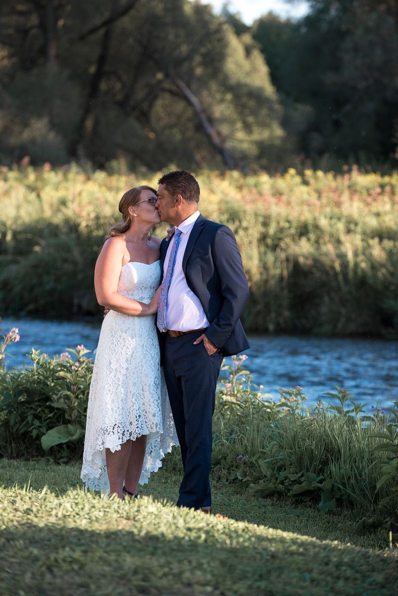Bride and groom kissing by a river, she in white lace dress, he in a blue suit.