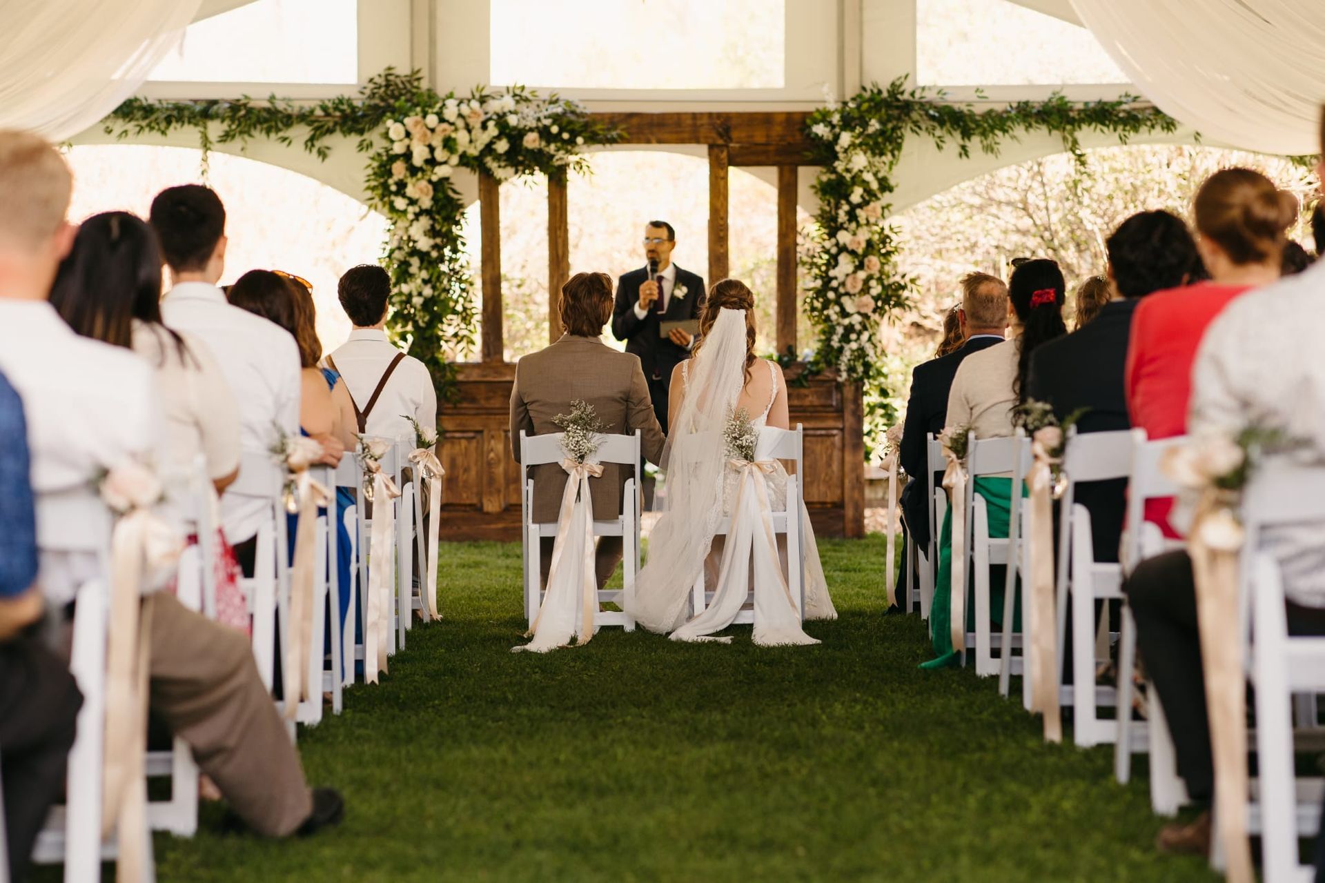 Wedding ceremony in a tent, with guests seated on white chairs facing the officiant.