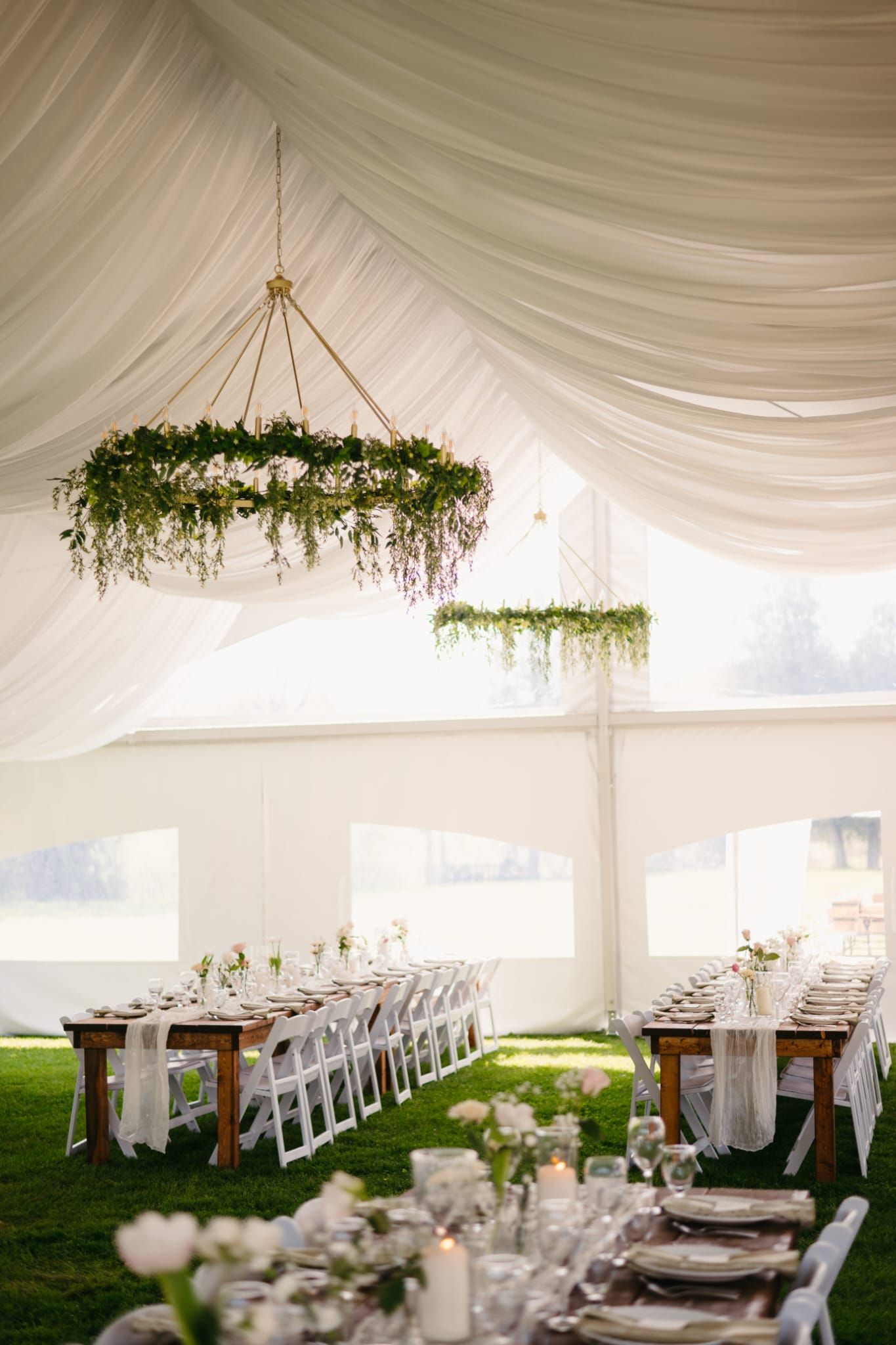 Elegant white wedding reception tent with draped ceiling, hanging greenery, and long decorated tables along a grassy aisle