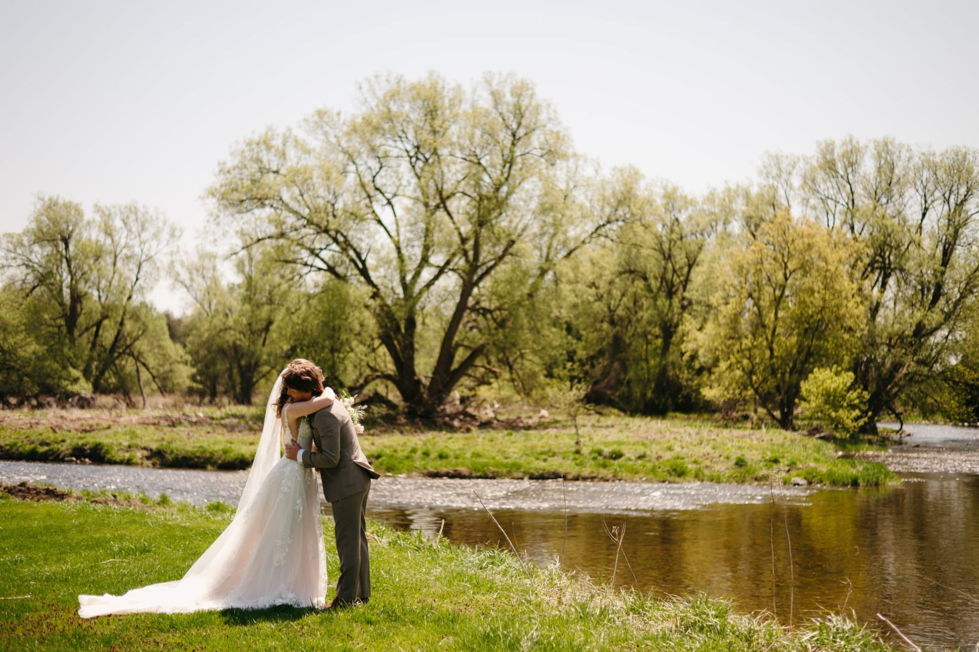 Bride and groom embrace by a pond in a green park with blooming trees
