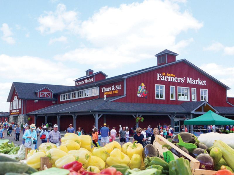 Farmers' Market building with red exterior, produce in foreground, and people shopping outdoors.