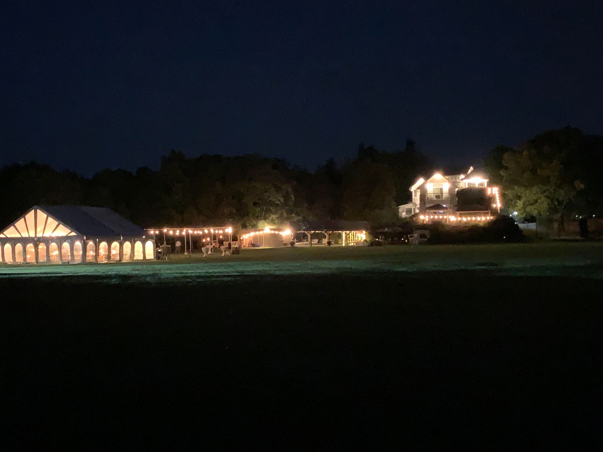 Night view of a venue lit up with string lights, including a tent, outdoor seating, and a house.