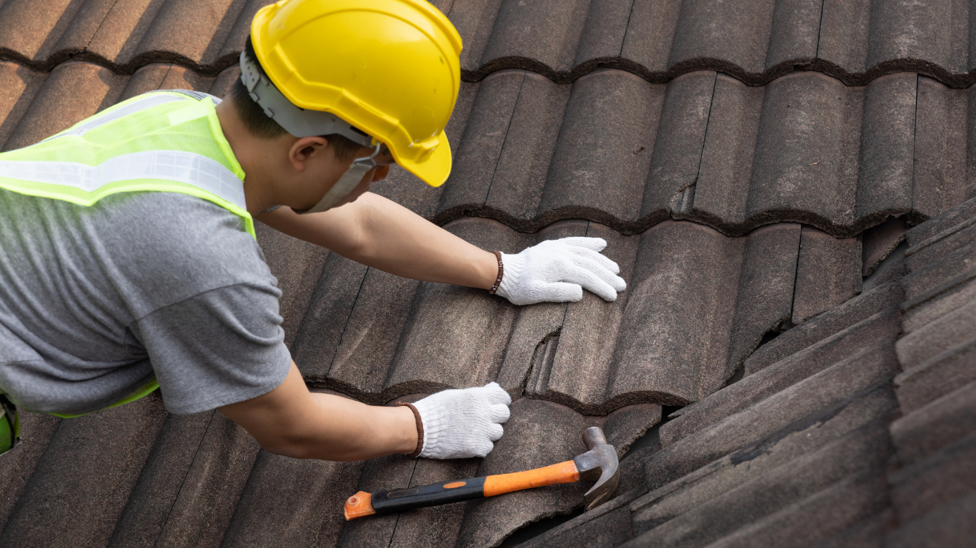 Roofer in yellow hard hat and gloves repairs tiled roof with a hammer.