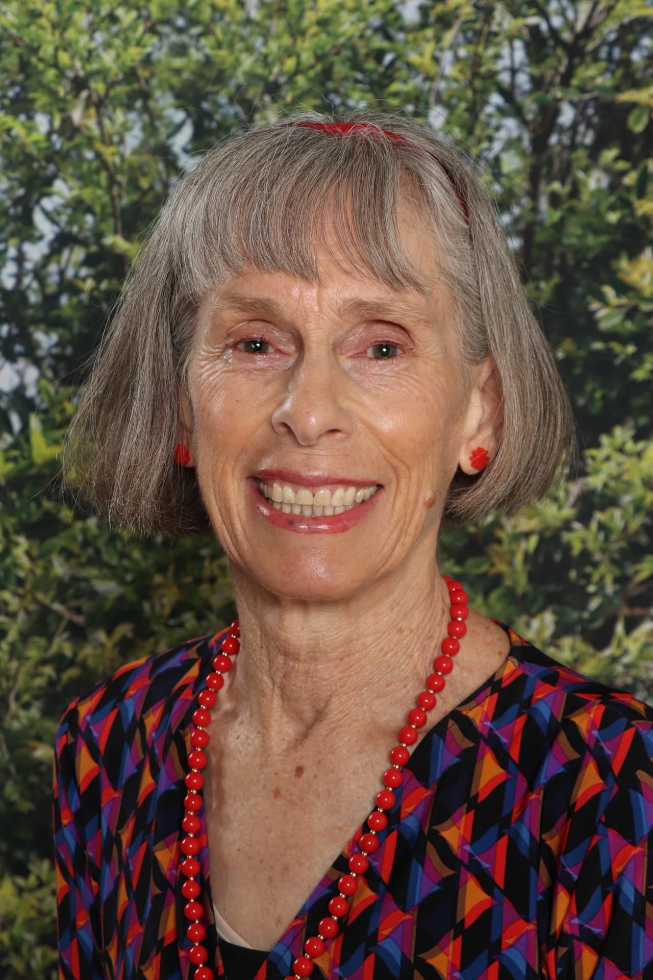 Woman with short gray hair, wearing a yellow floral blouse and red bead necklace, smiles in front of red wall with artwork.