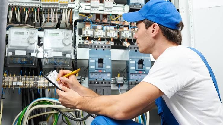 Technician in blue cap inspecting electrical control panel with tools in industrial setting