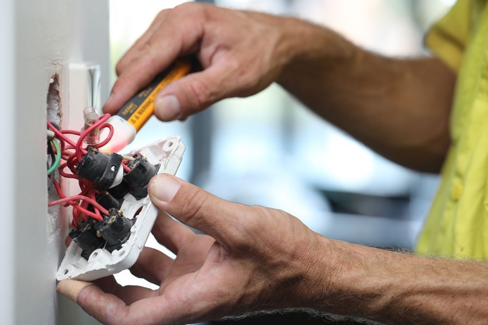 Hands using a screwdriver to repair exposed electrical wiring in a wall outlet.
